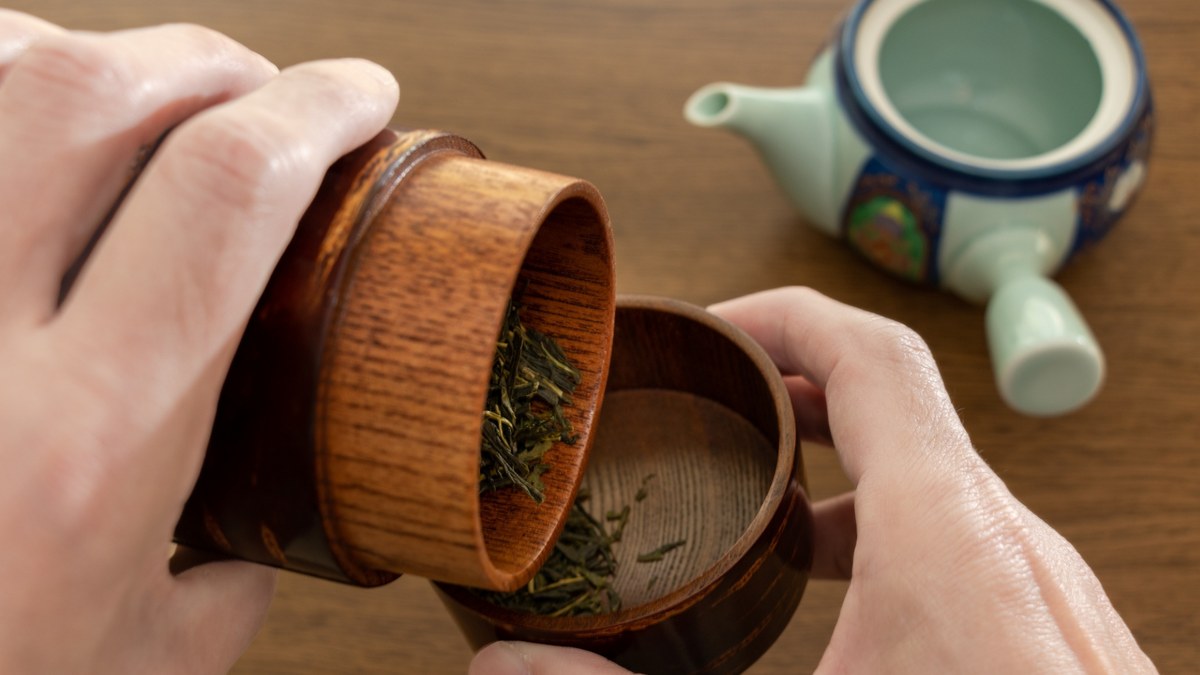 Hands lifting the inner lid from a Japanese cherry-bark tea caddy, with a kyusu behind