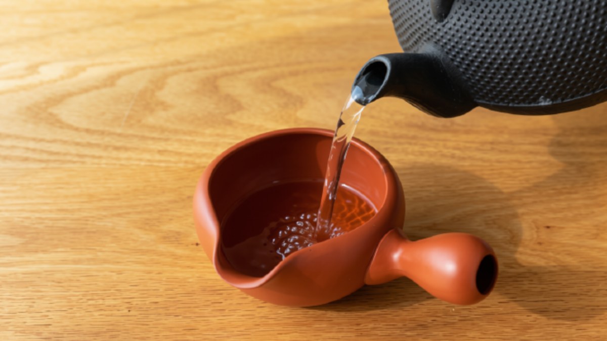 An iron kettle pouring hot water into a clay yuzamashi on a wooden table