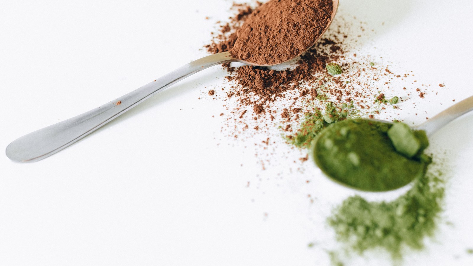 A spoon of brown hojicha powder beside a spoon of green matcha powder