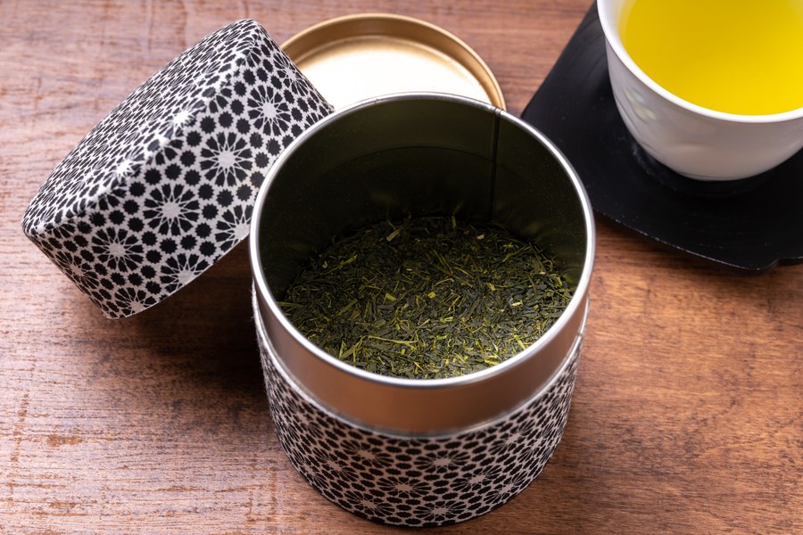 A Japanese tea caddy with its lid off, sencha inside, beside a cup of green tea