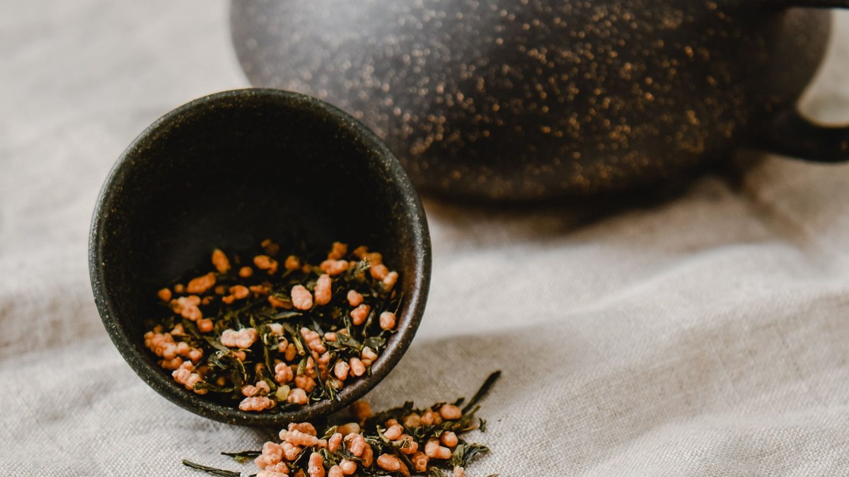 Loose genmaicha leaves — green tea blended with roasted brown rice — spilling from a black cup beside a teapot