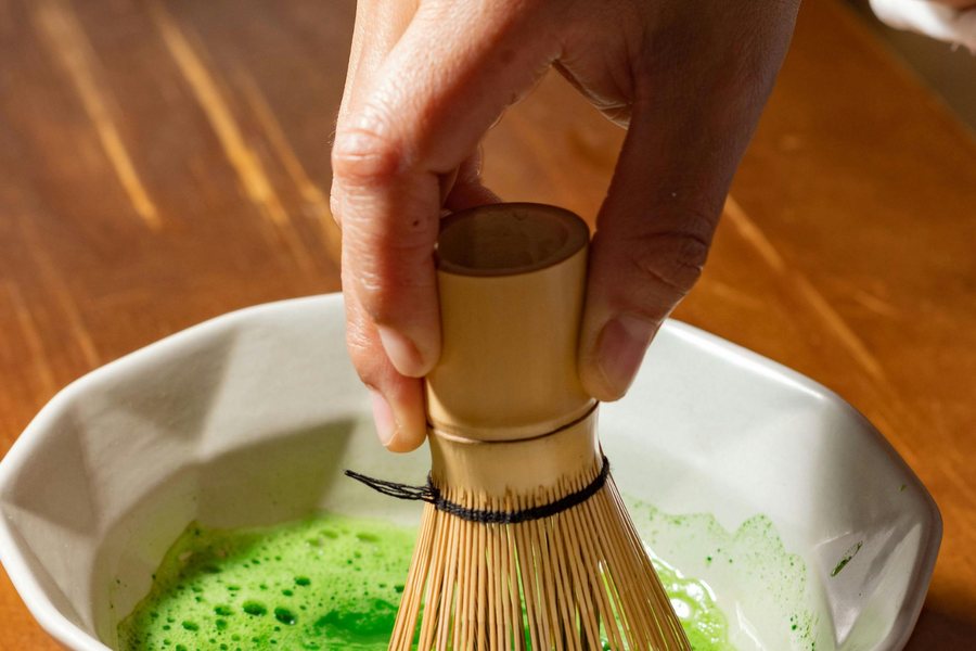 A bamboo whisk frothing bright green matcha in a ceramic bowl