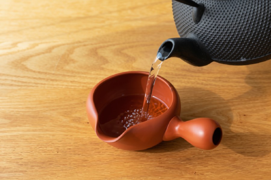 An iron kettle pouring hot water into a red clay yuzamashi with a pouring lip