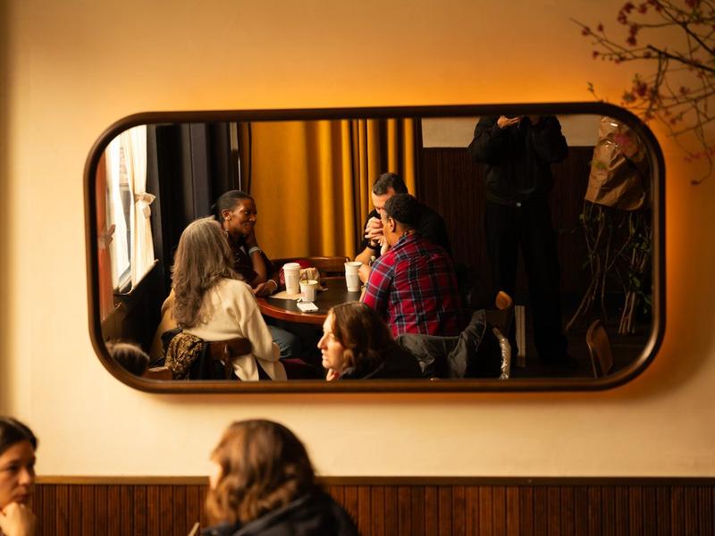 A mirror on the wall reflecting people talking over coffee at a round table